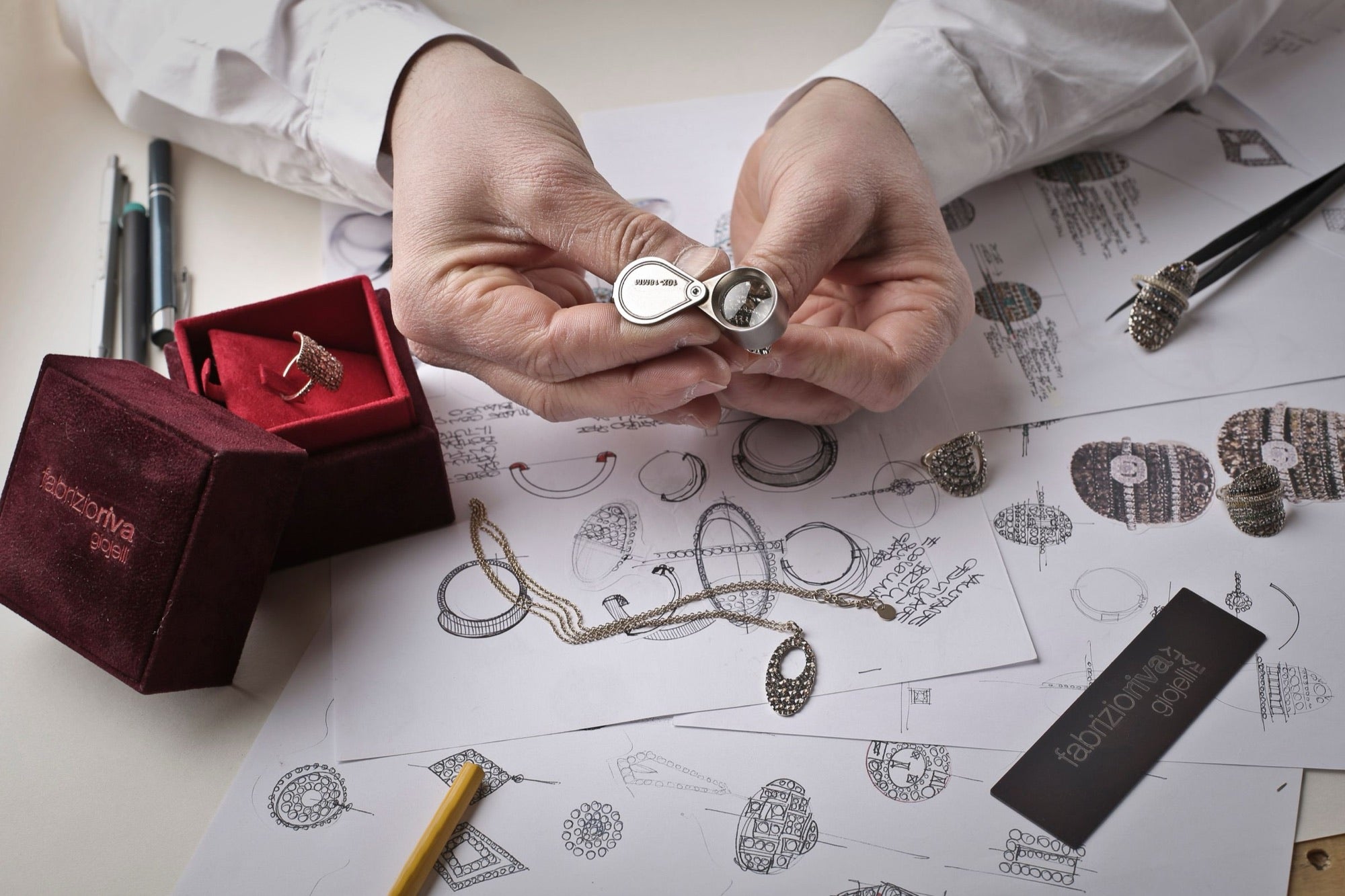 A jeweller inspecting a ring above sketches at a workbench.